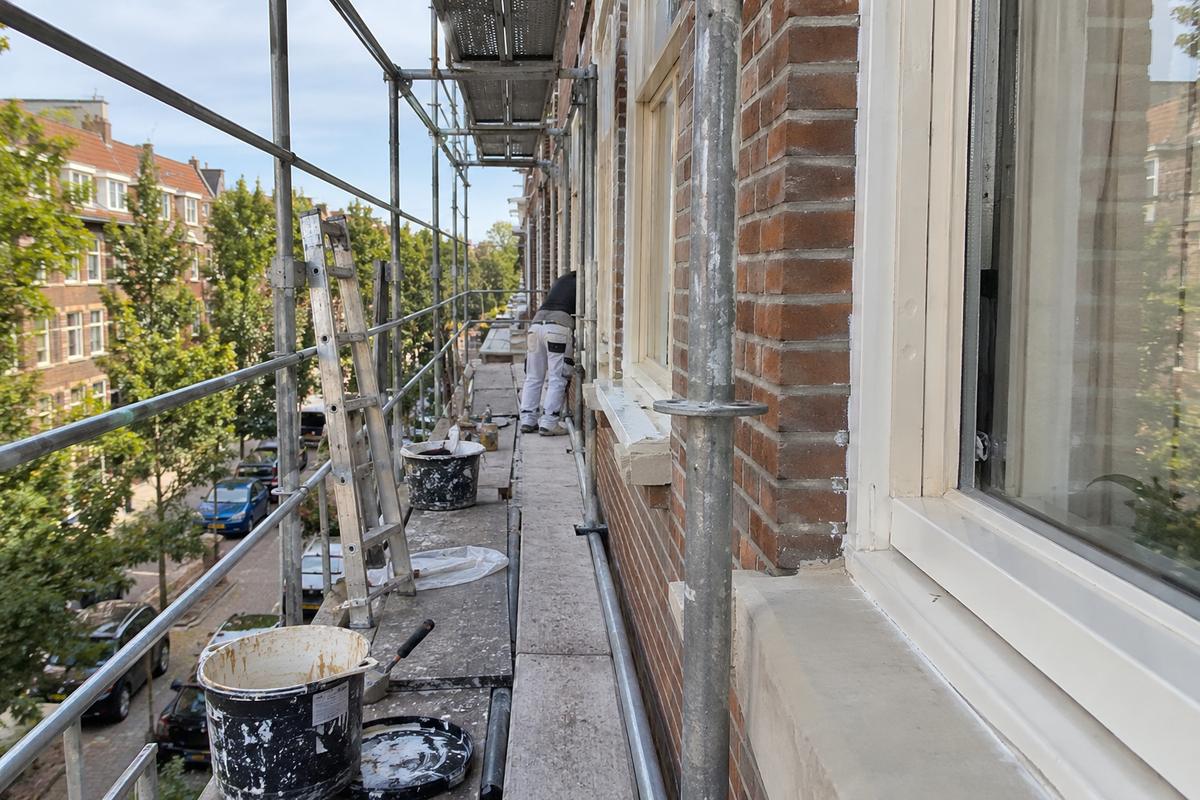 Steiger langs een bakstenen woonblok tijdens schilderwerk aan de kozijnen, met emmers en een ladder