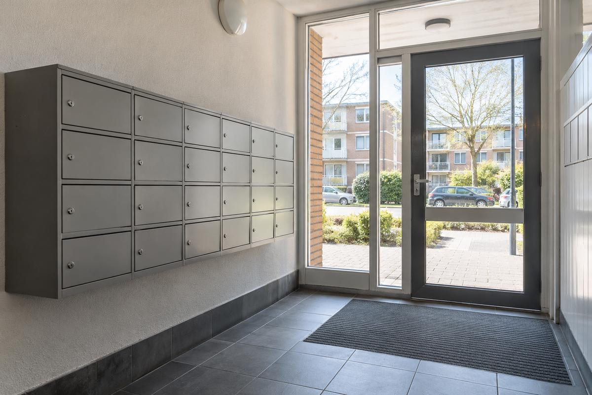 Entree van een appartementengebouw met een wand vol brievenbussen, een deurmat en een glazen voordeur met uitzicht op de straat