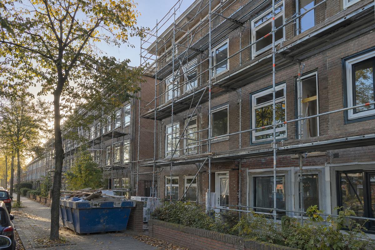 Steigers langs de bakstenen gevel van een portiekflat tijdens een renovatie, met deels geplaatste nieuwe kozijnen en een bouwcontainer aan de stoeprand