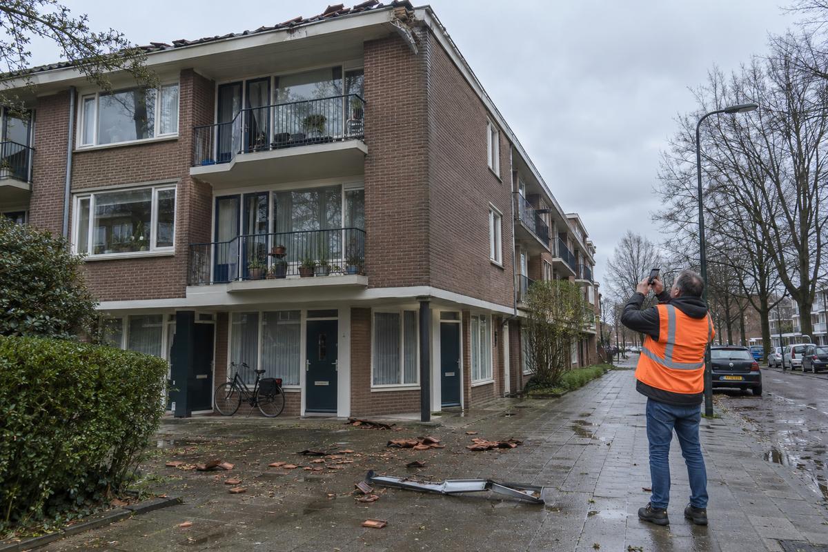 Bakstenen flat met balkons na een storm, met gebroken dakpannen en een losgeraakte dakgoot op de natte stoep en een man in een oranje hesje die de dakrand fotografeert