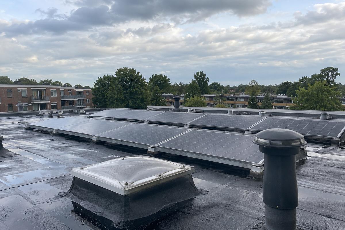 Plat dak van een bakstenen flat met rijen zonnepanelen, een daklicht en een ontluchtingspijp, met daken en bomen op de achtergrond